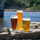A trio of refreshing wheat beers—American Wheat, Bavarian Hefeweizen, and Dunkelweizen—displayed in distinct glasses atop a riverside rock with a scenic bridge and tree-lined background, ideal for promoting a wheat beer homebrew variety pack.
