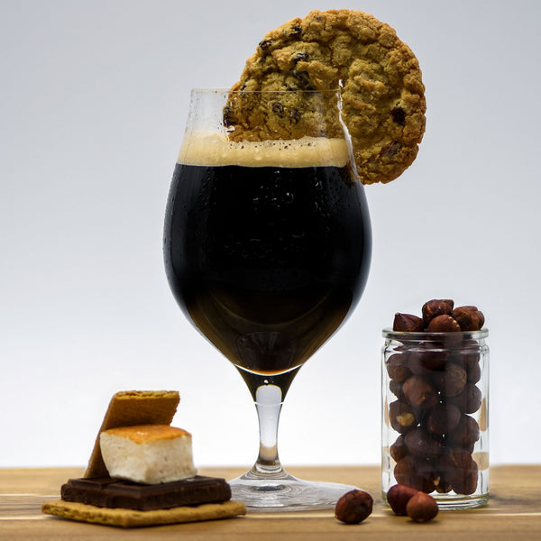 A glass of Sweet Tooth Pastry Stout with a cookie wedge beside a glass filled with hazelnuts and a fully-assembled smores