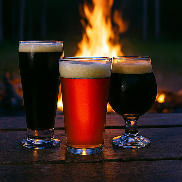 Image of 3 Beers Two dark ales and one red ale on a table in front of a campfire in the woods. 
